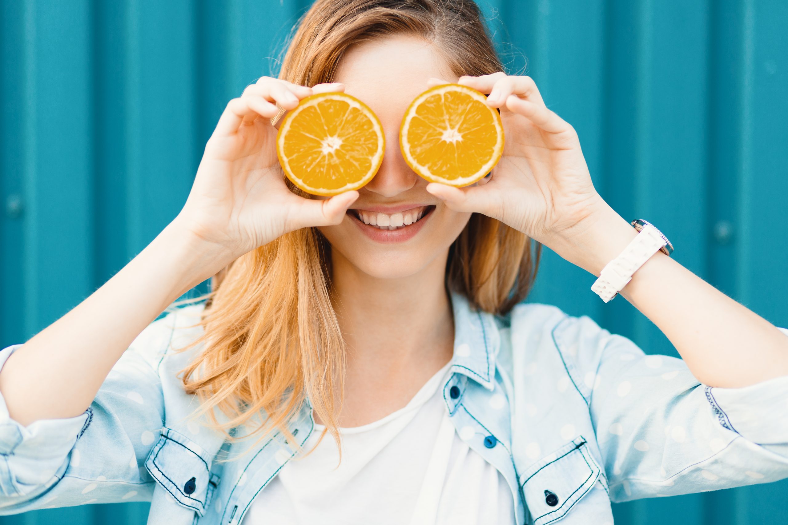 Carefree young beautiful girl using two halfs of oranges instead of binocular over her eyes on blue background.